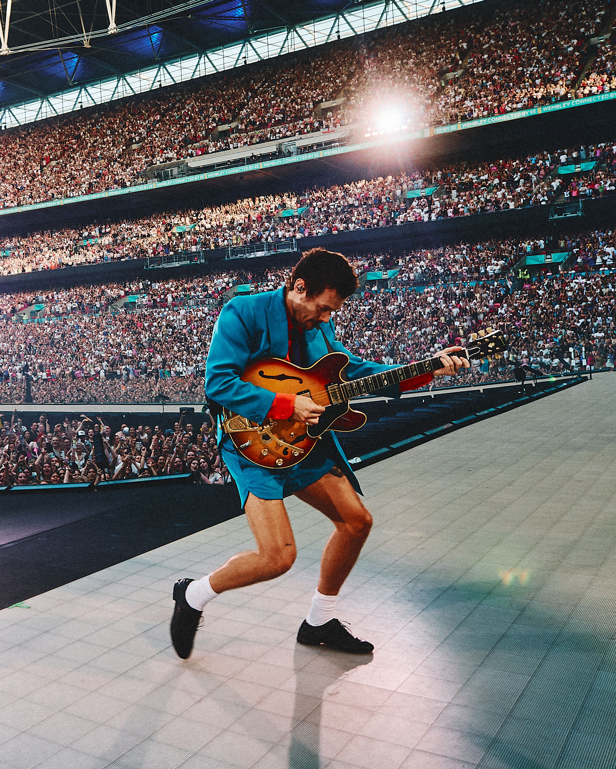 Harry Styles playing guitar on stage at Wembley with fans behind him